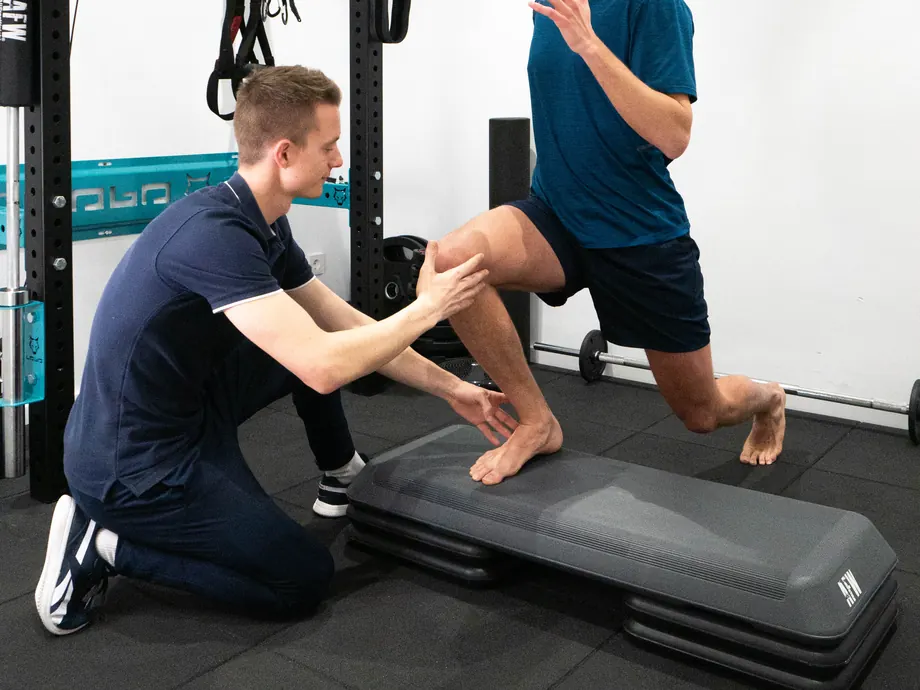 Un fisioterapeuta, de rodillas, sujeta la rodilla de un deportista que hace una zancada sobre un step en la sala de ejercicio de Activefisio